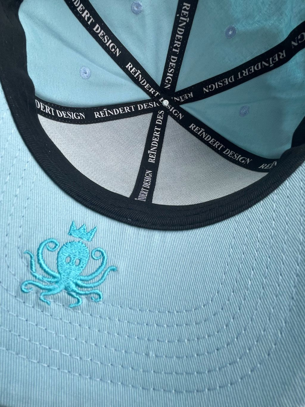 Vue de dessous de la casquette bleu turquoise en plan serré sur le logo pieuvre, poulpe du dessous de la visière